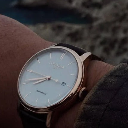 A close-up of a LOCMAN wristwatch with a rose gold case, white dial, and brown leather strap worn on a person's wrist against a blurred outdoor background.