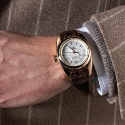 A close-up of a wrist wearing a vintage-style watch with a white dial, gold-toned case, and dark brown leather strap, worn under a brown pinstripe jacket.